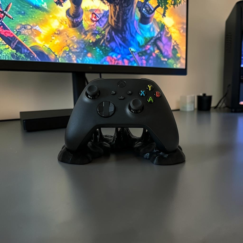 Black Xbox controller displayed on a black, uniquely shaped stand with a sticky effect, on a white desk next to a lamp.