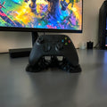 Black Xbox controller displayed on a black, uniquely shaped stand with a sticky effect, on a white desk next to a lamp.