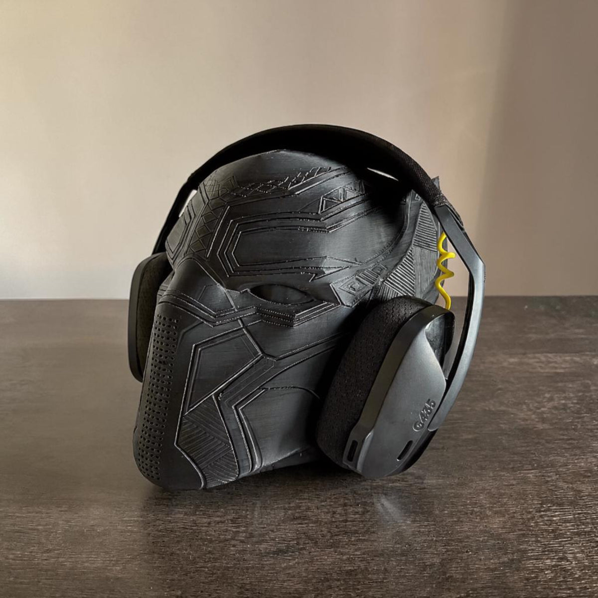 Black Panther mask headphone stand by Magicland 3D. Black 3D printed stand with intricate mask details, holding black headphones.