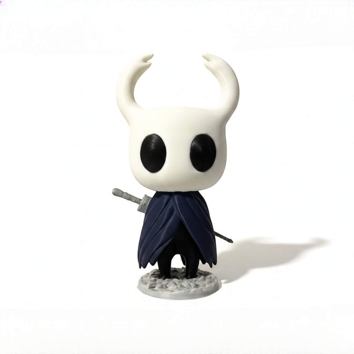 Magicland 3D Hollow Knight figürü, beyaz boynuzlu, siyah gözlü ve lacivert pelerinli, kılıcı elinde tutuyor.