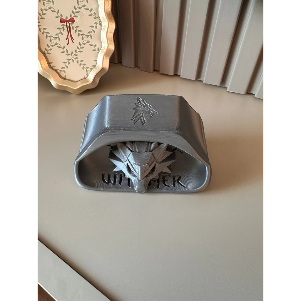 Magicland 3D Witcher temalı gri 3D baskı telefon tutucu stant, üzerinde kurt başı sembolü ve Witcher yazısı bulunur.