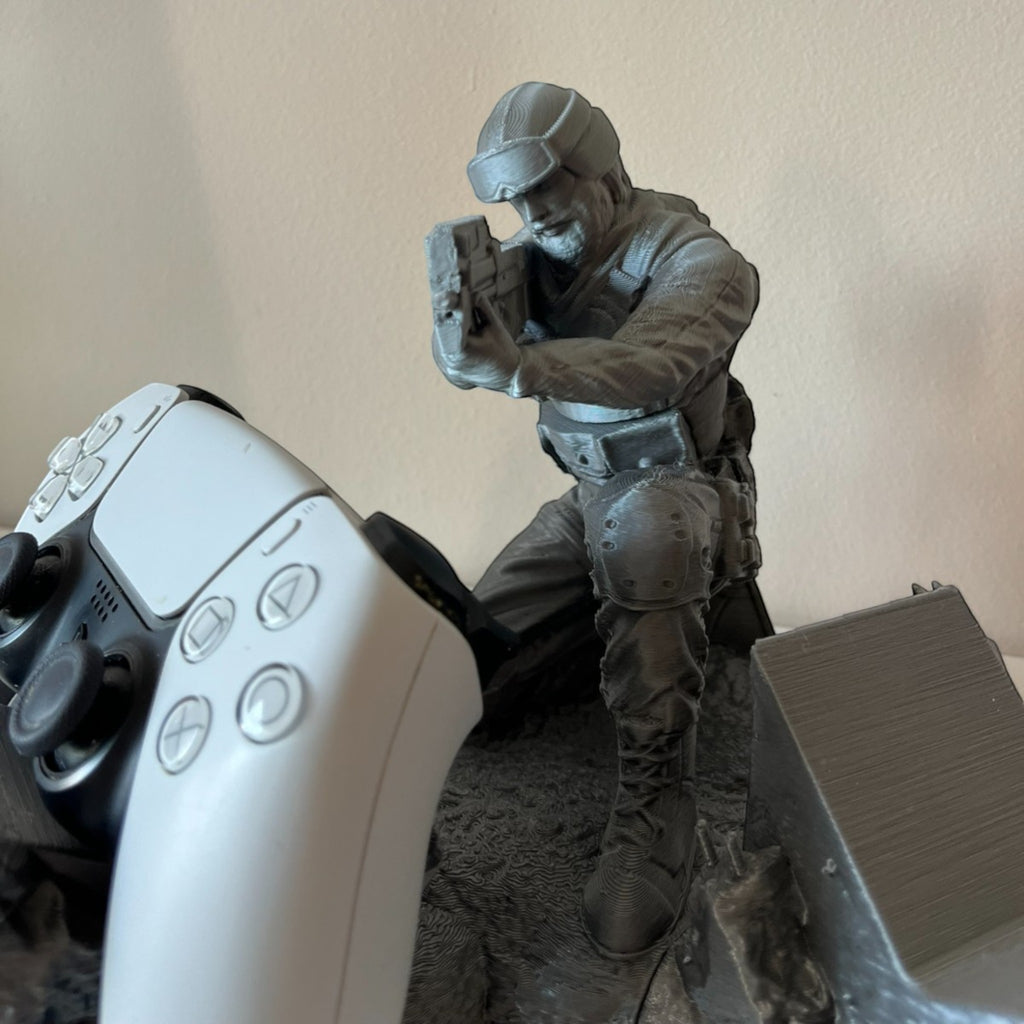 Magicland 3D Asker figure controller stand, grey, detailed soldier figurine holding a gun, next to a white PS5 controller.