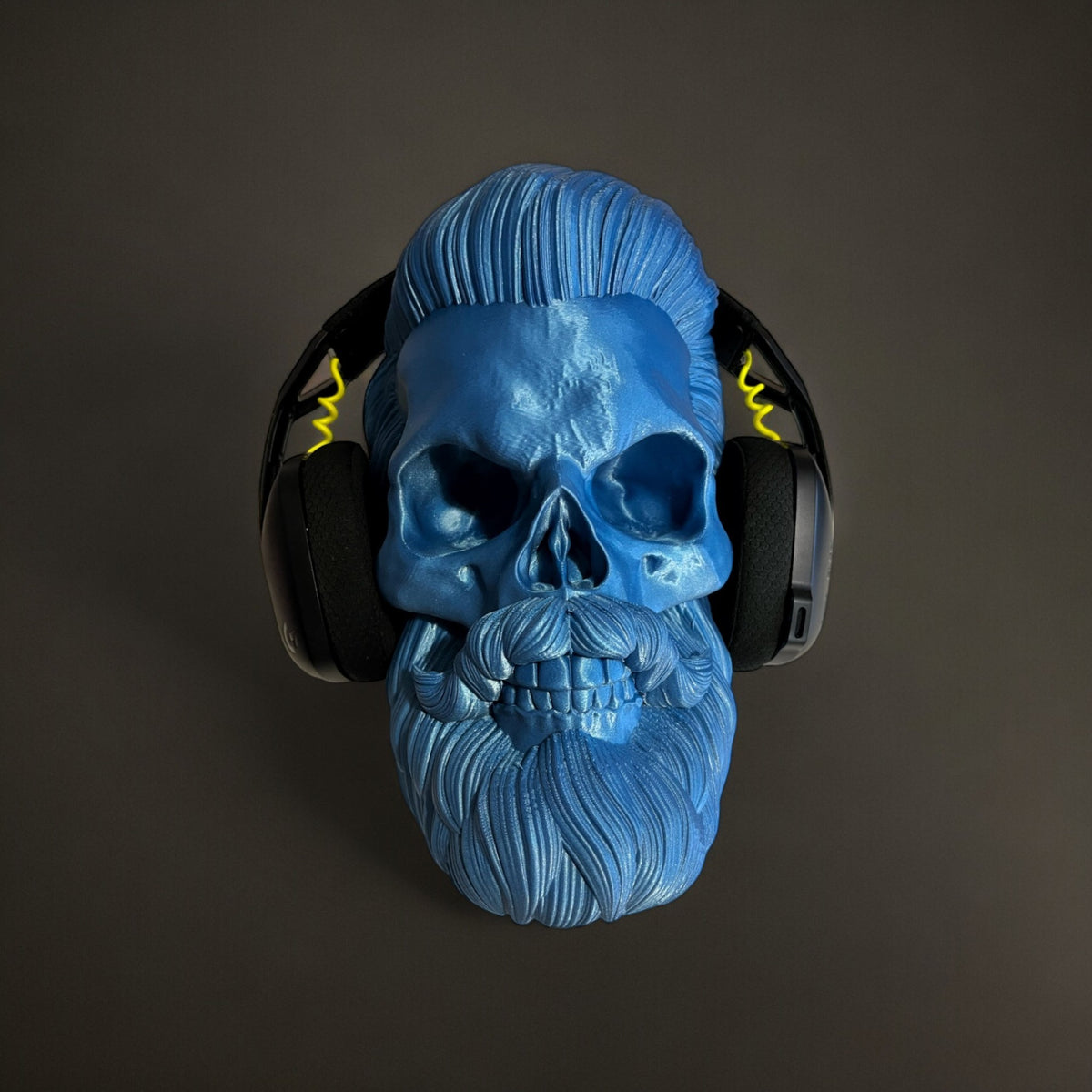Blue 3D printed skull headphone stand with a beard and slicked-back hair, holding black headphones with yellow accents.