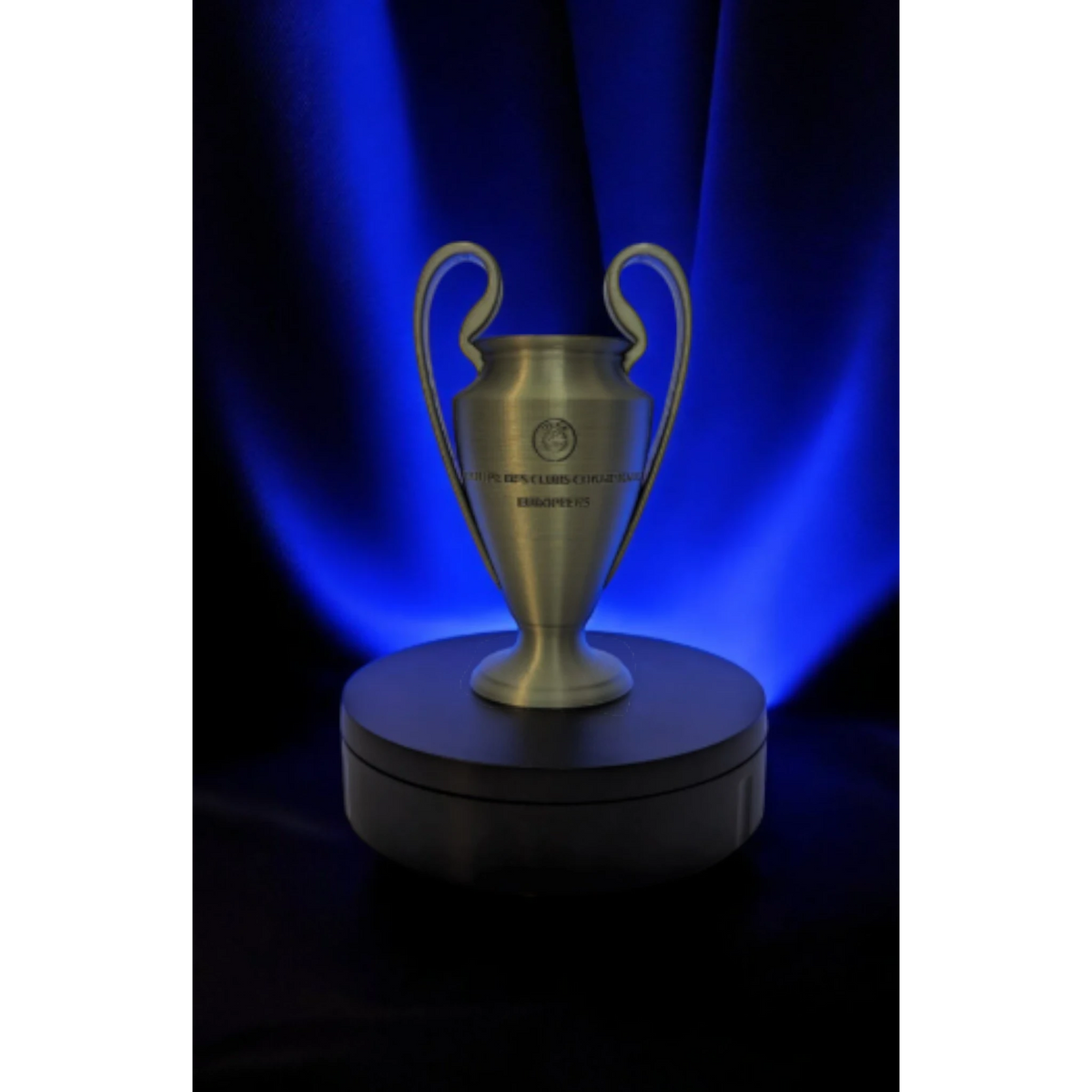 Magicland 3D replica of the Champions League trophy, a metallic gold cup with two handles on a rotating black stand against a blue backdrop.